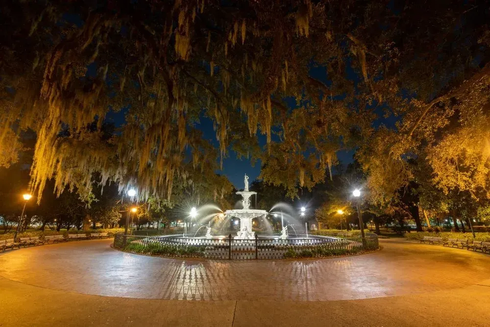 Nighttime park fountain with lit water jets, surrounded by trees and streetlights