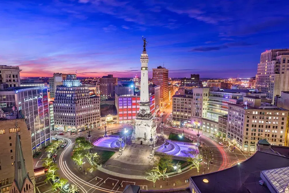 Dusk view of a city square with a tall monument, colorful lights, and surrounding buildings under a blue sky