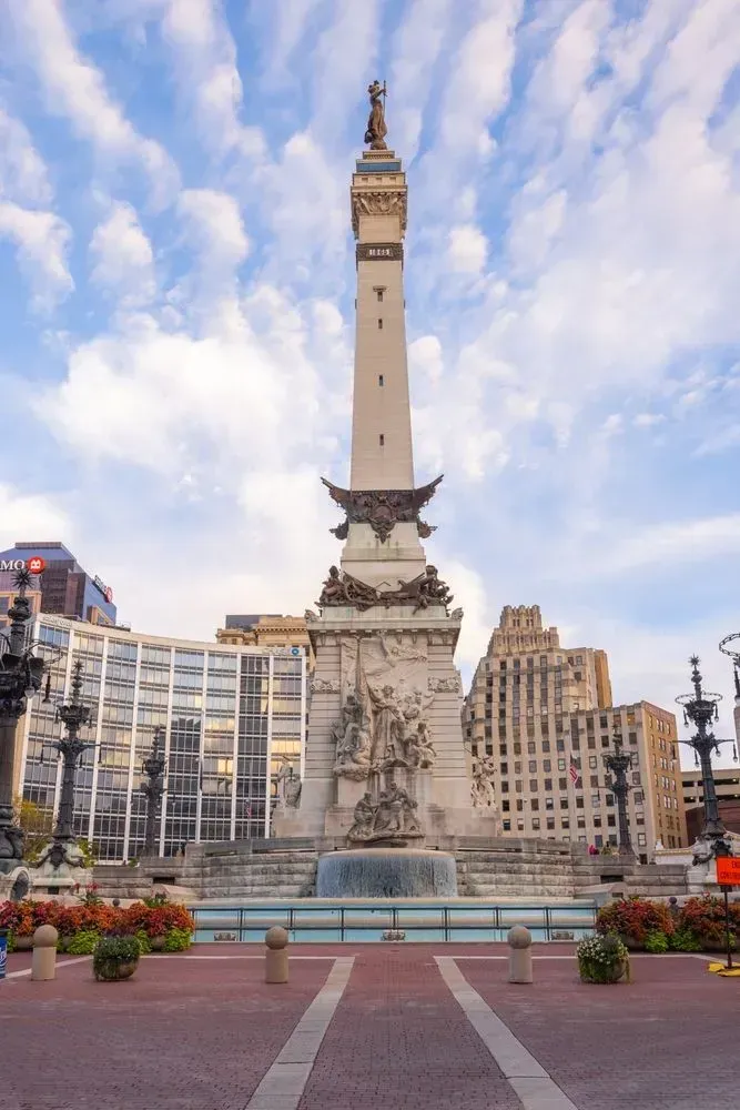 Monument in a city square with a tall white column, framed by modern buildings and flower beds under a cloudy sky