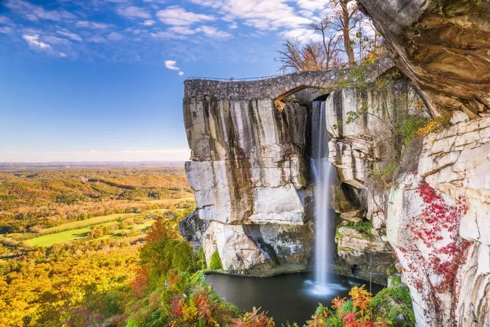 Waterfall plunging from a sheer cliff into a pool, overlooking a bright autumn valley under a blue sky.