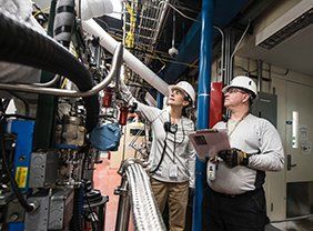 Two engineers in hard hats inspect industrial equipment in a factory room