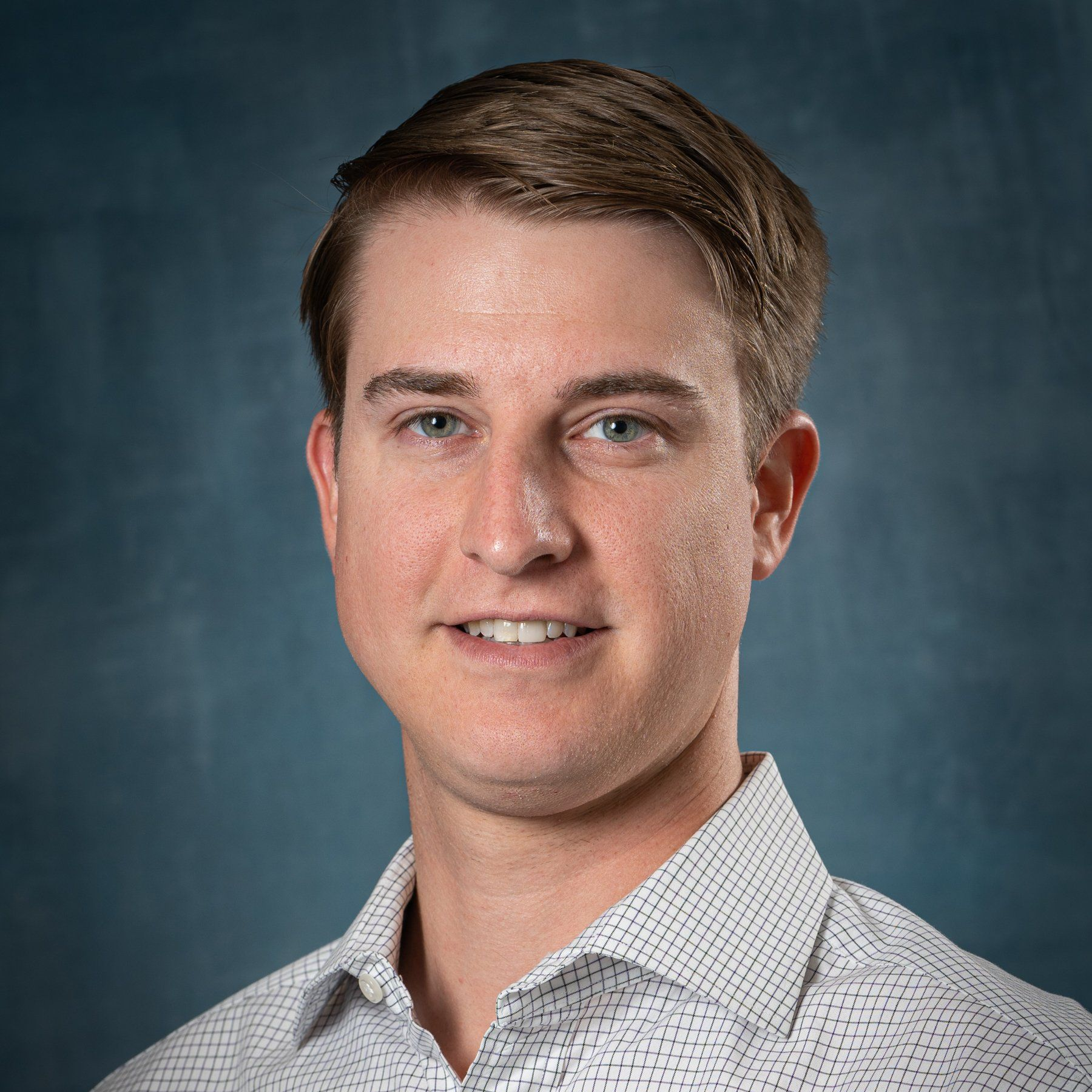 Studio headshot of a person in a light gray patterned shirt against a blue-gray background
