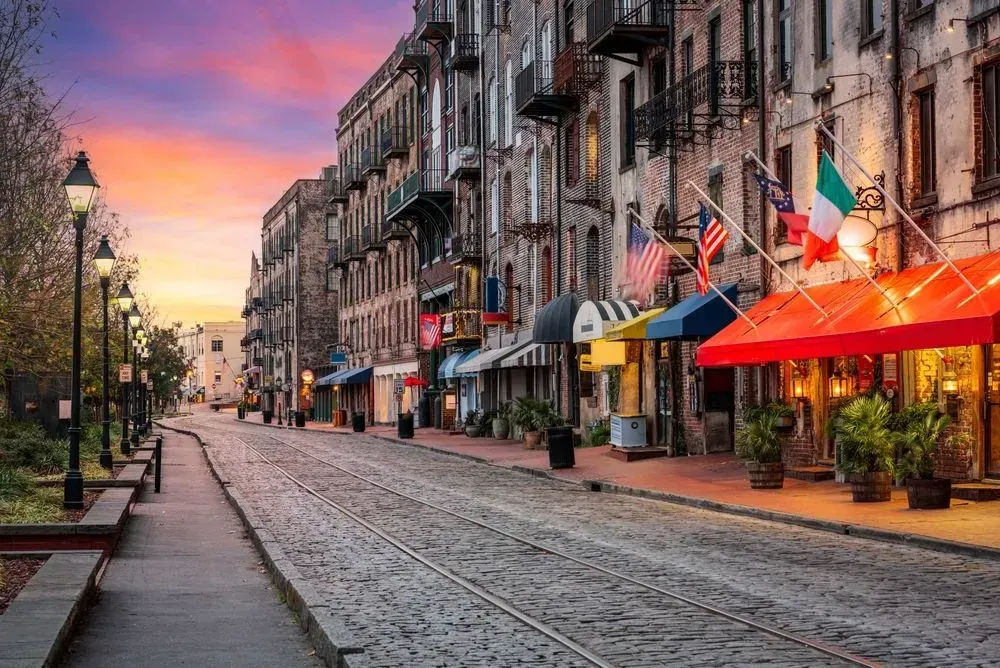 Quiet cobblestone street at sunset with historic buildings, benches, and colorful awnings glowing under a pink sky