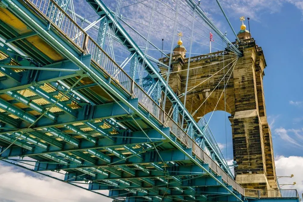 Blue glass walkway beneath a stone suspension bridge tower against a clear sky