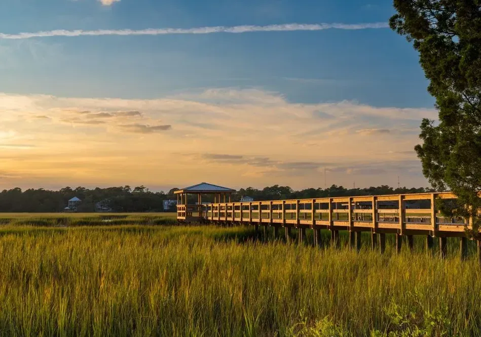 Wooden boardwalk through marsh grass at sunset, leading to a lakeside pavilion under a golden sky
