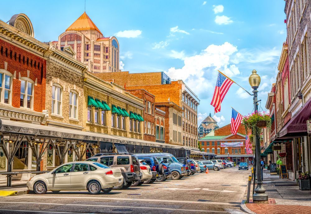 Historic downtown street with parked cars, brick storefronts, and American flags under a bright blue sky