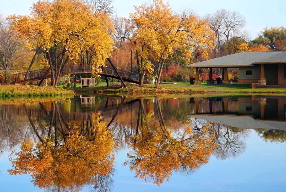 Autumn trees reflected in a calm lake beside a lodge and dock, with golden foliage and clear blue sky.