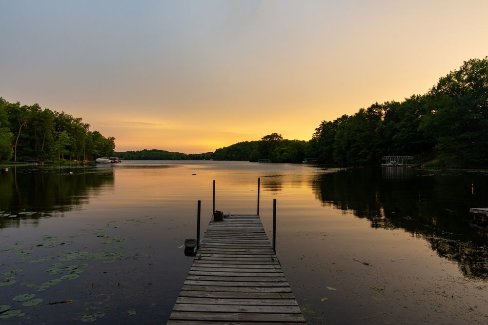 Wooden dock extending into a calm lake at sunset, framed by dark trees and a glowing orange sky