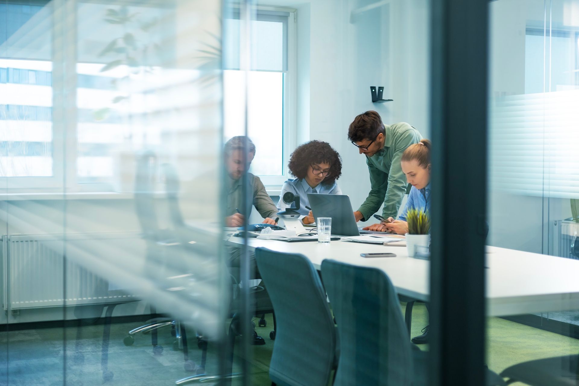 People in a glass meeting room gathered around a laptop at a white conference table.