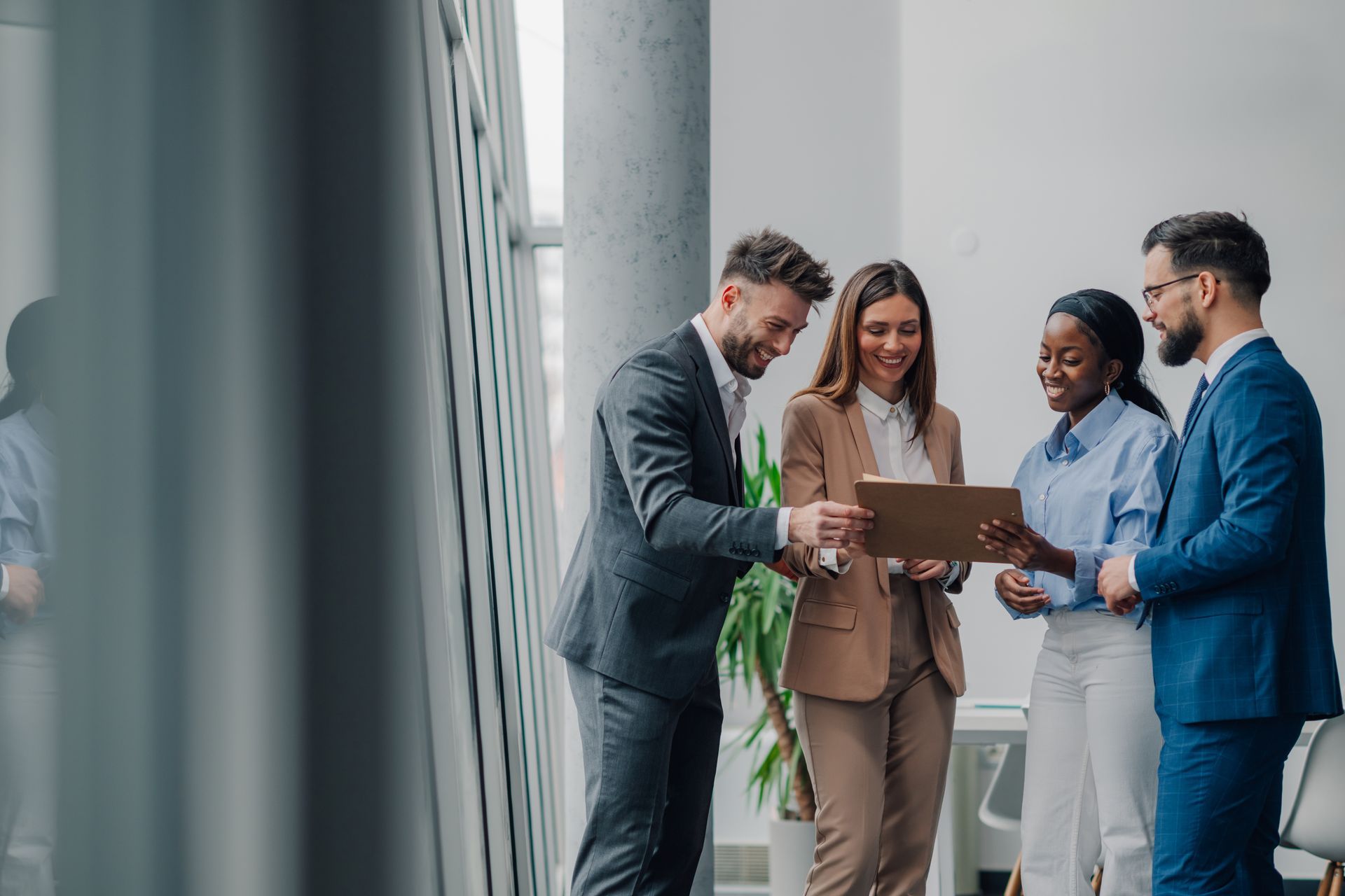 Four businesspeople standing by a window, reviewing a folder and smiling in a bright office