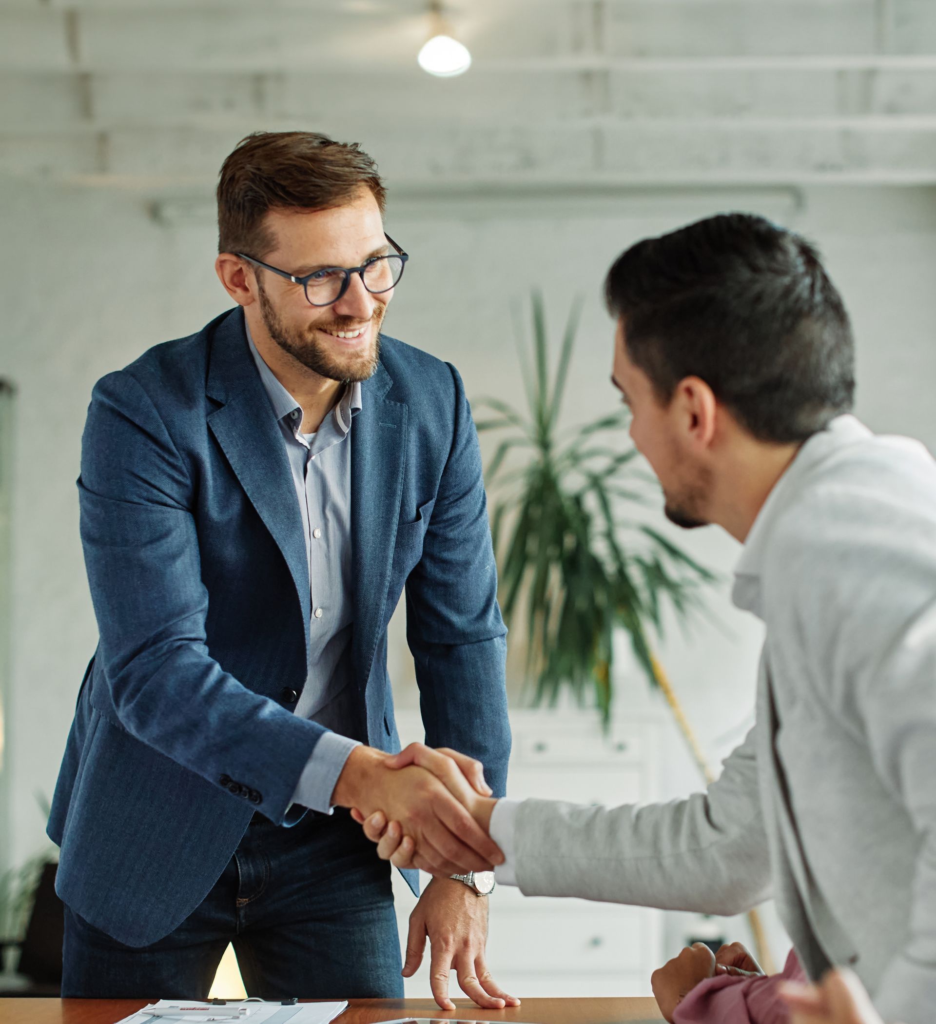 Two men shake hands across a desk in a bright office, one standing in a blue blazer.