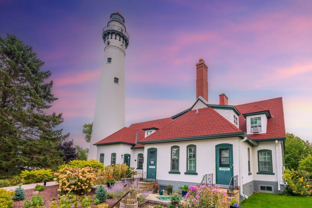 White lighthouse and red-roofed house beside a colorful garden at sunset