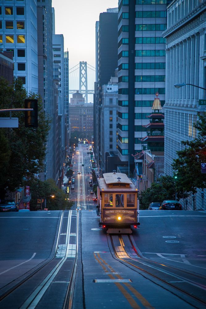 Cable car descending a steep city street at dusk, framed by tall buildings and the Bay Bridge in the distance