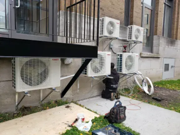 Une personne installe des unités de climatisation sur des supports le long du mur d'un bâtiment.