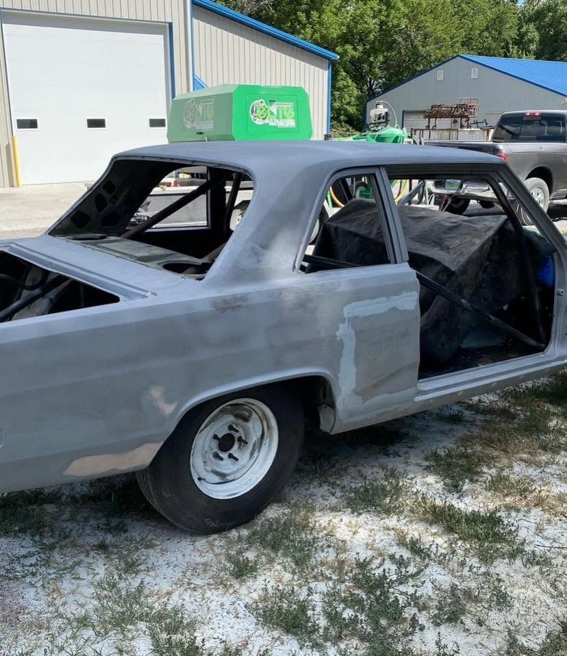 Gray unfinished car body on white rims parked outdoors near a garage, with open windows and no doors.