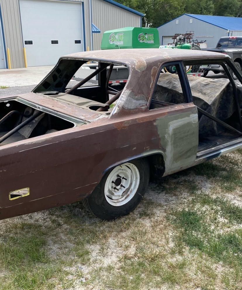 Rusty classic car shell in a driveway, maroon and gray primer, missing windows and interior panels.