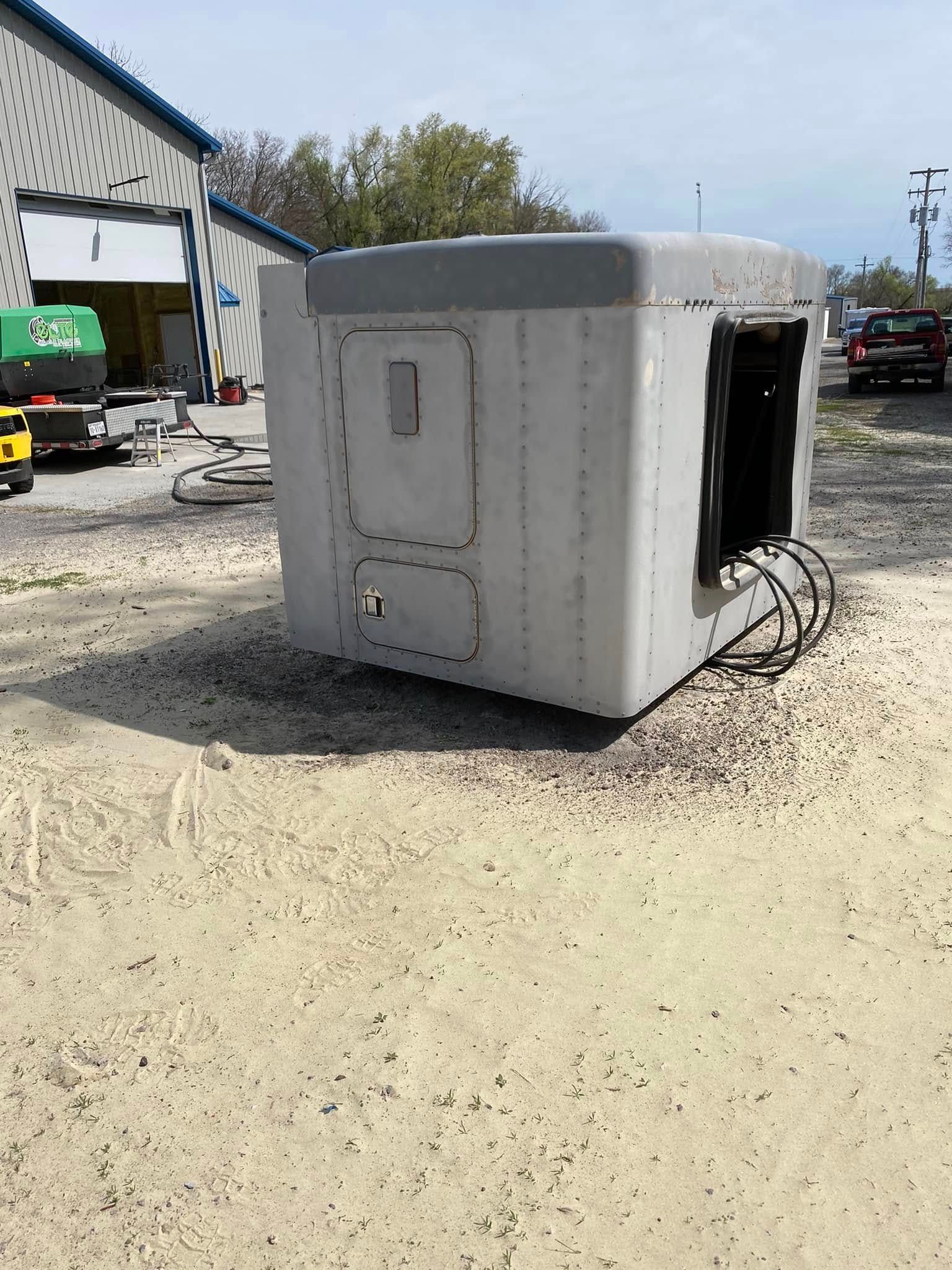 Large gray industrial container or equipment box on a gravel lot beside a workshop, with cables trailing out.