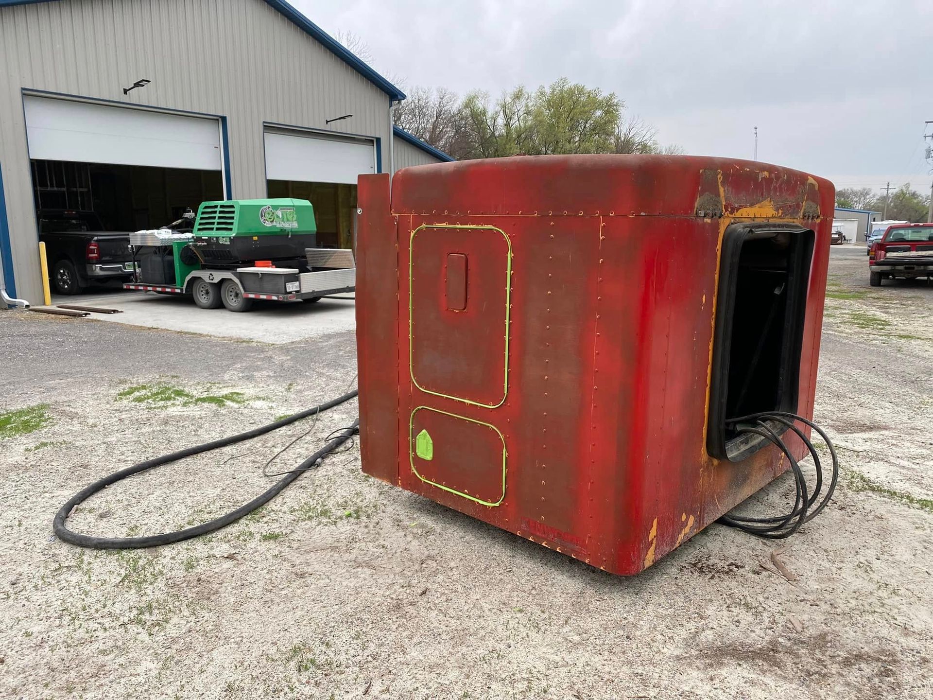 Red shipping container with open doorway sitting on gravel beside a warehouse and green truck