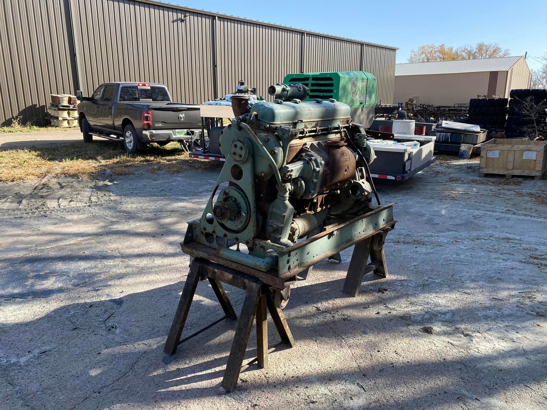 Old green engine on a stand in a gravel lot with trucks and shipping containers behind it