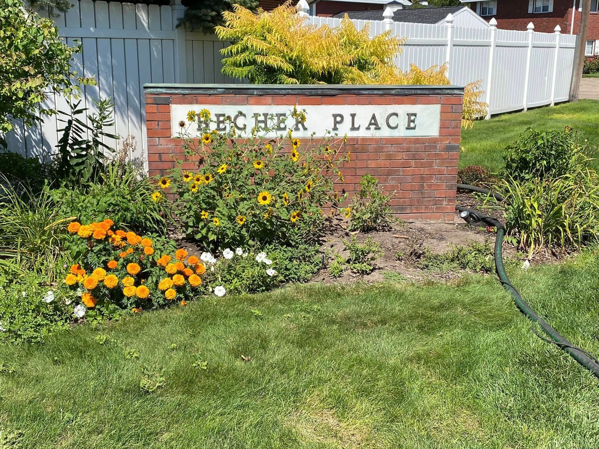 Brick sign reading “BEECHER PLACE” in a flower bed with orange and yellow blooms and a white fence behind it