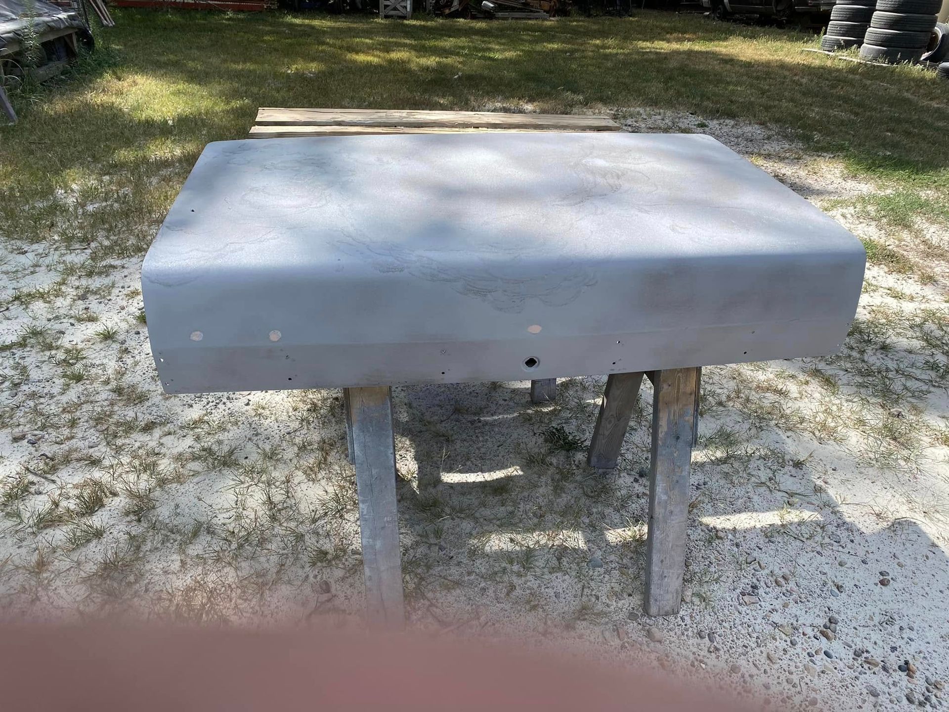 Gray outdoor table or bench on a sandy ground in a grassy yard, with two metal legs and trees in the background