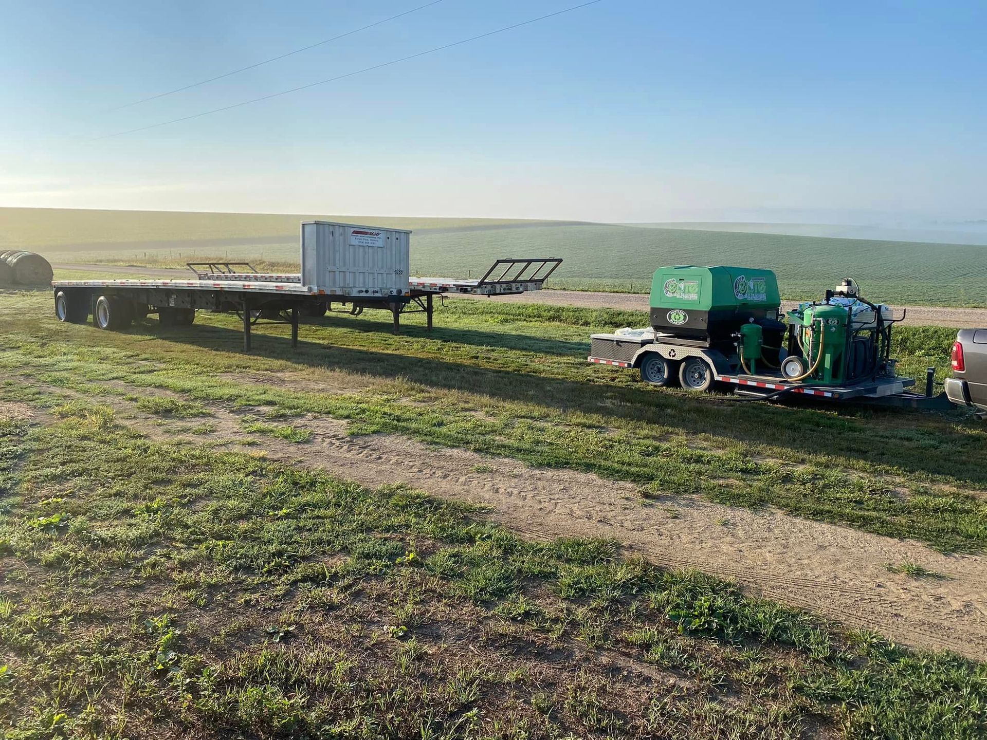 Two utility trailers on a grassy hill at sunset, with equipment and a person working nearby