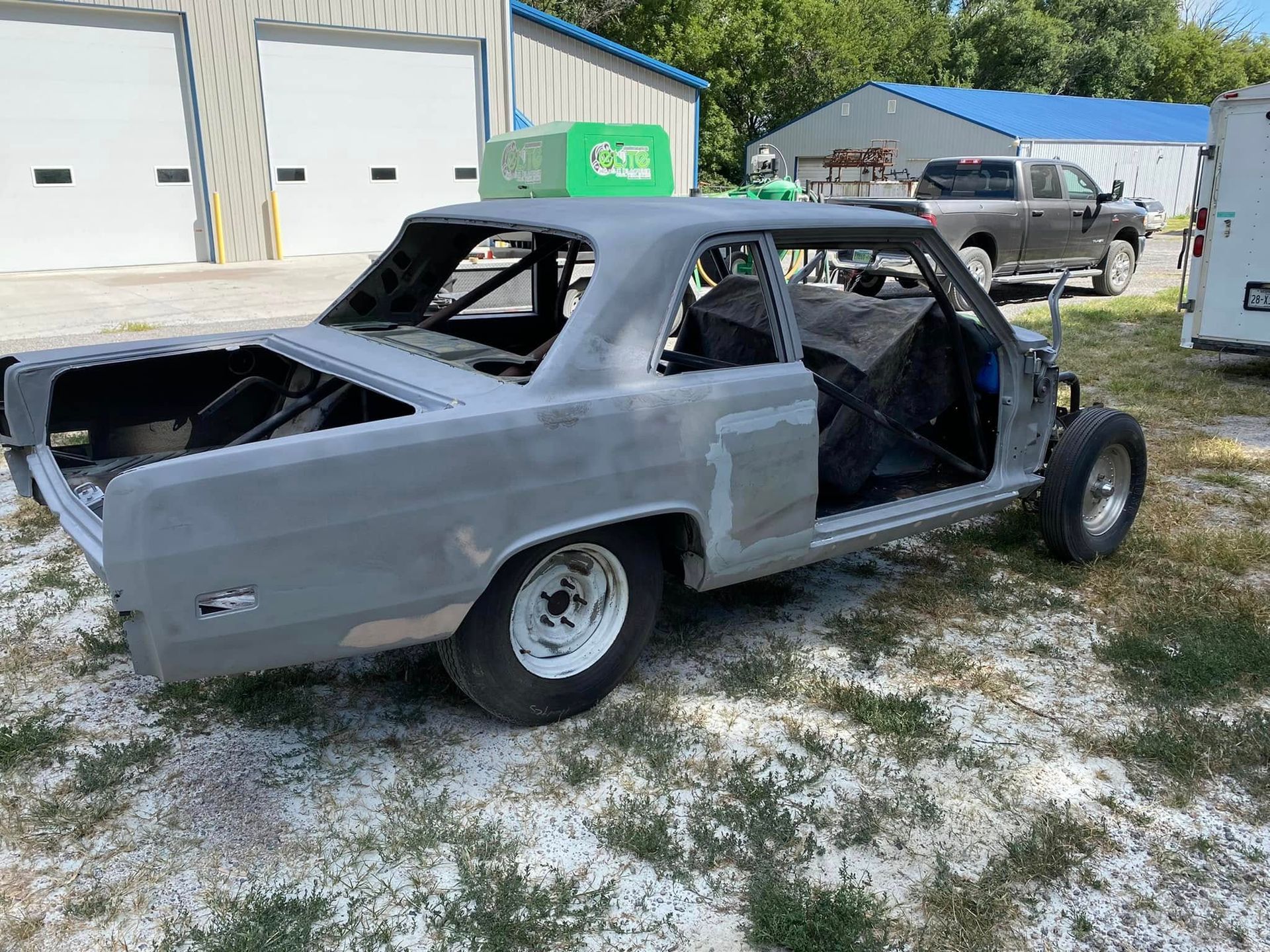Gray stripped car body on wheels parked outdoors beside a garage, with no doors or windows.