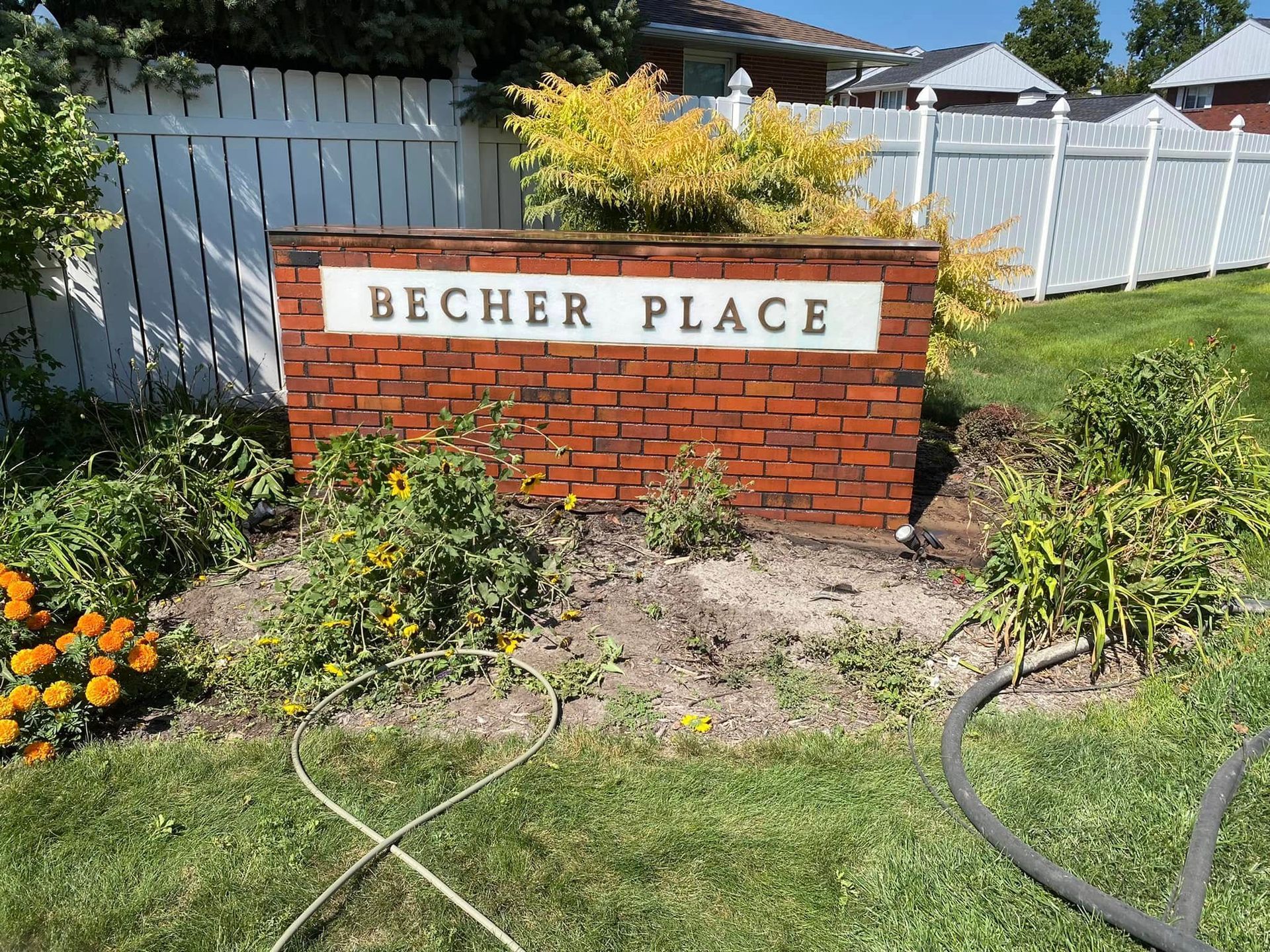 Brick sign reading “BECHER PLACE” in a garden with flowers, grass, and a white fence.