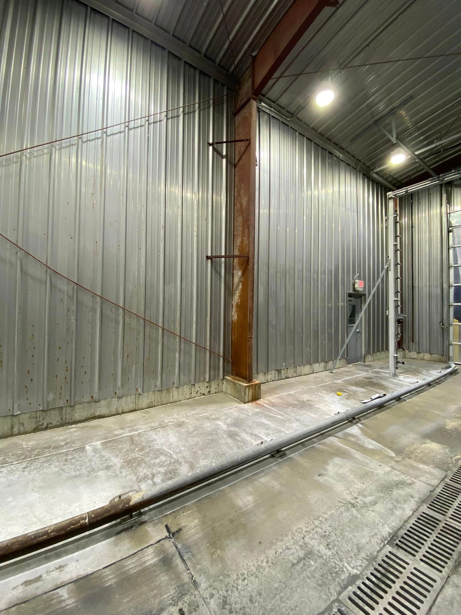 Corridor with corrugated metal walls, a wooden beam, and wet floor drains under bright overhead lights