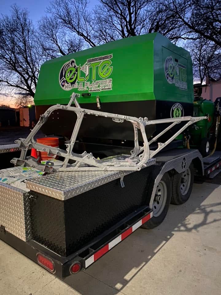 Green off-road buggy frame on a trailer at dusk, with a black storage box and orange cones nearby.