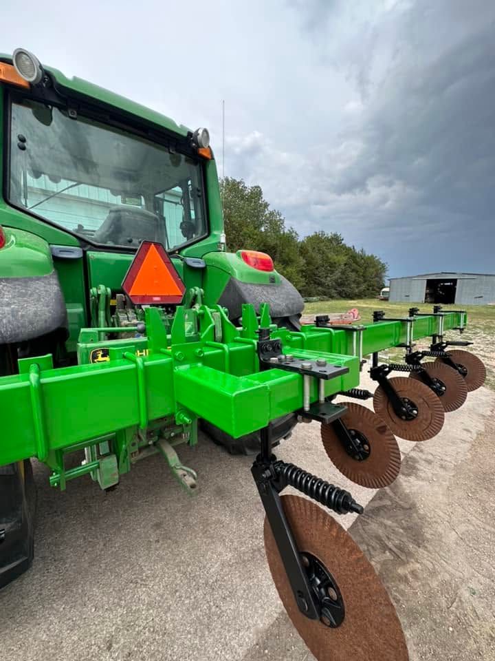 Green tractor with attached brown disc harrow parked on a gravel lot under cloudy sky