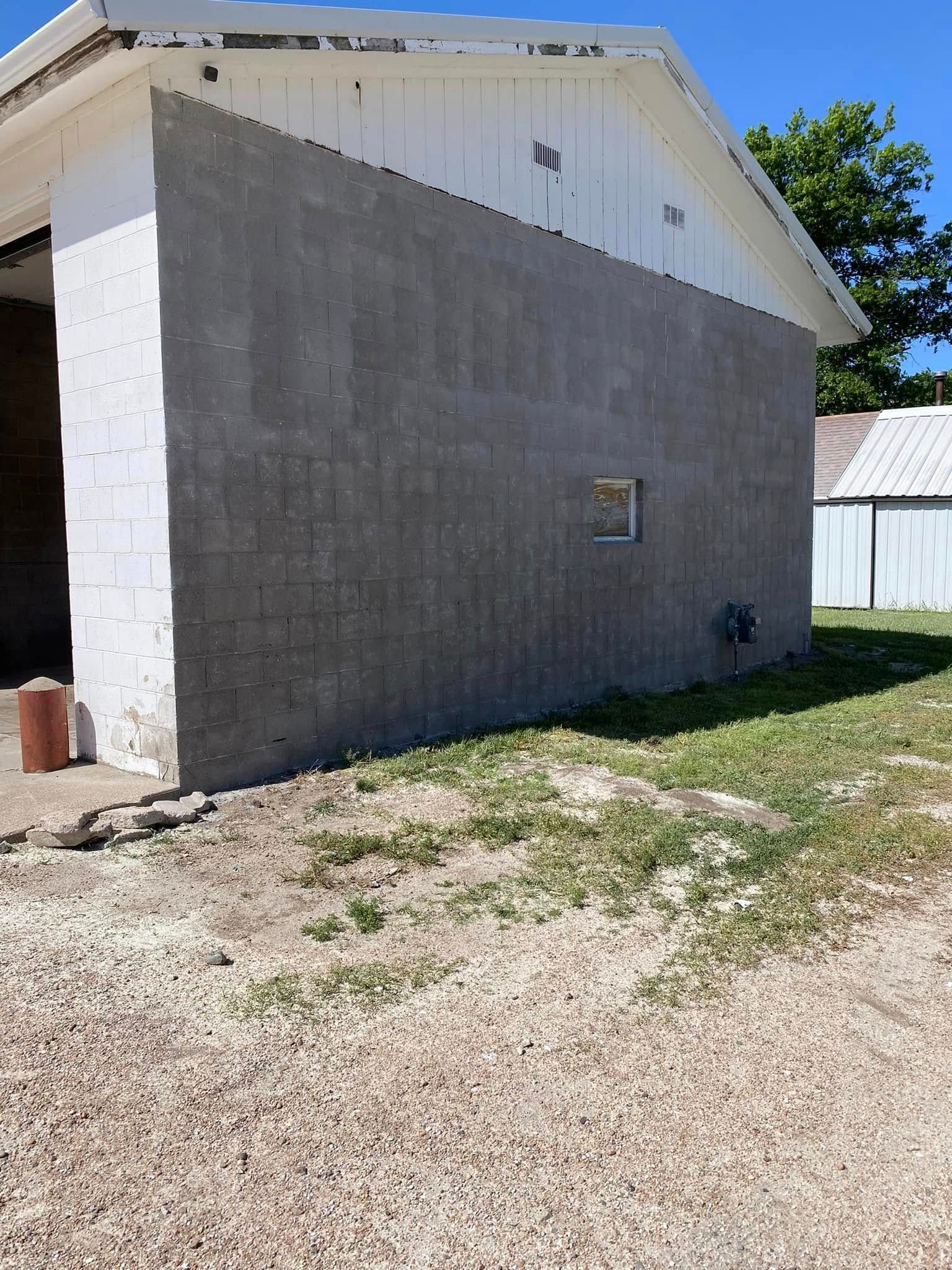 Gray cinder block building with white upper siding, gravel yard, and trees in bright sunlight