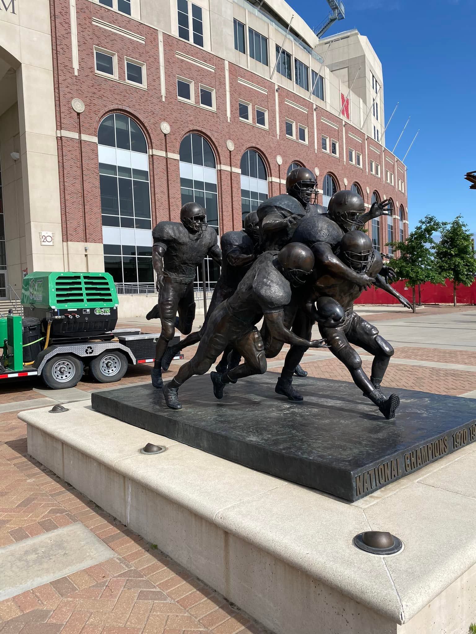 Bronze football tackle statue on a stone pedestal outside a brick stadium under a blue sky