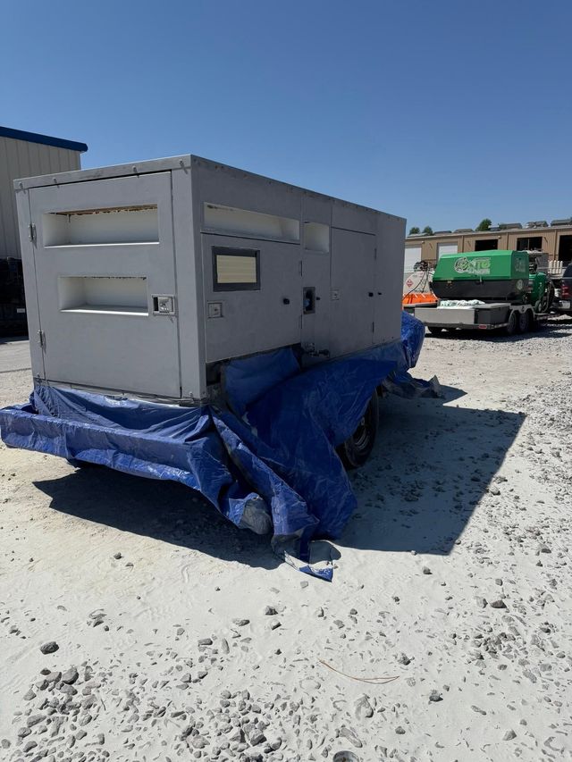 Gray industrial machine on blue tarp in a gravel yard, with a green truck in the background.