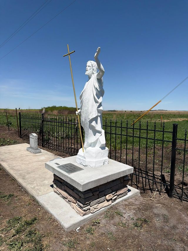White roadside religious statue with cross on a concrete pedestal beside a fence in a rural field
