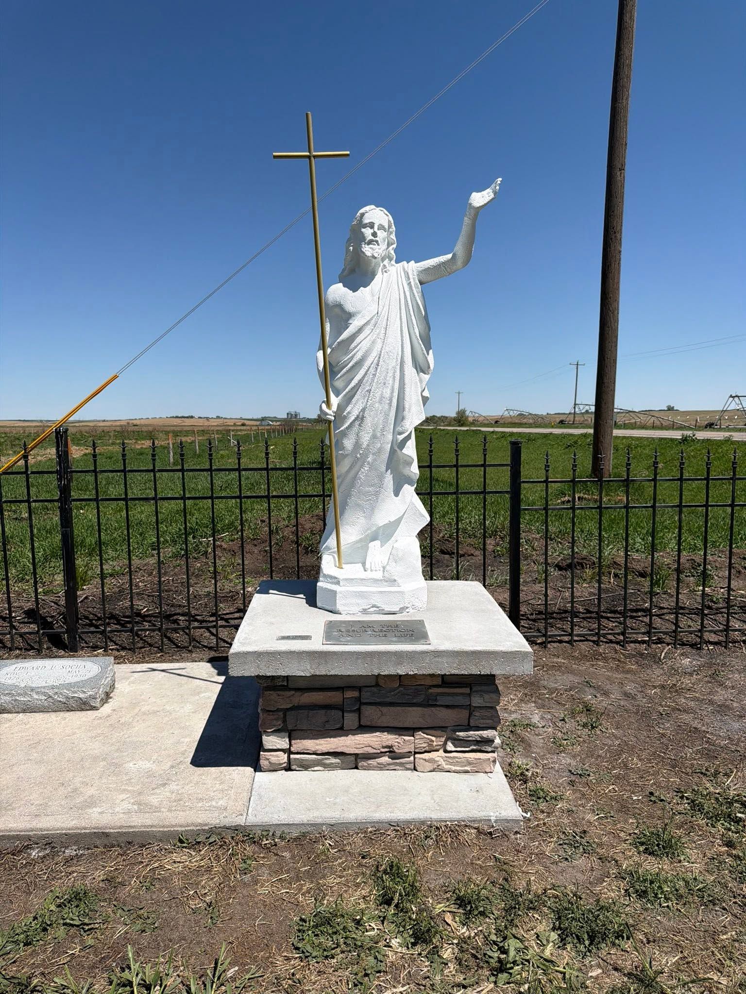 White statue of a saint with raised arm beside a cross in a fenced outdoor area under a blue sky