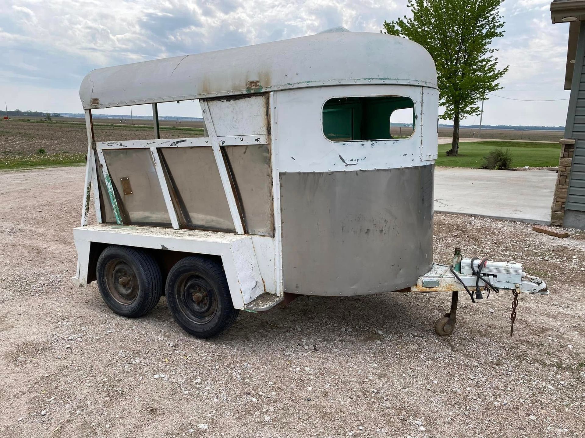 Small white horse trailer with black wheels parked on gravel, shown in a rural field.