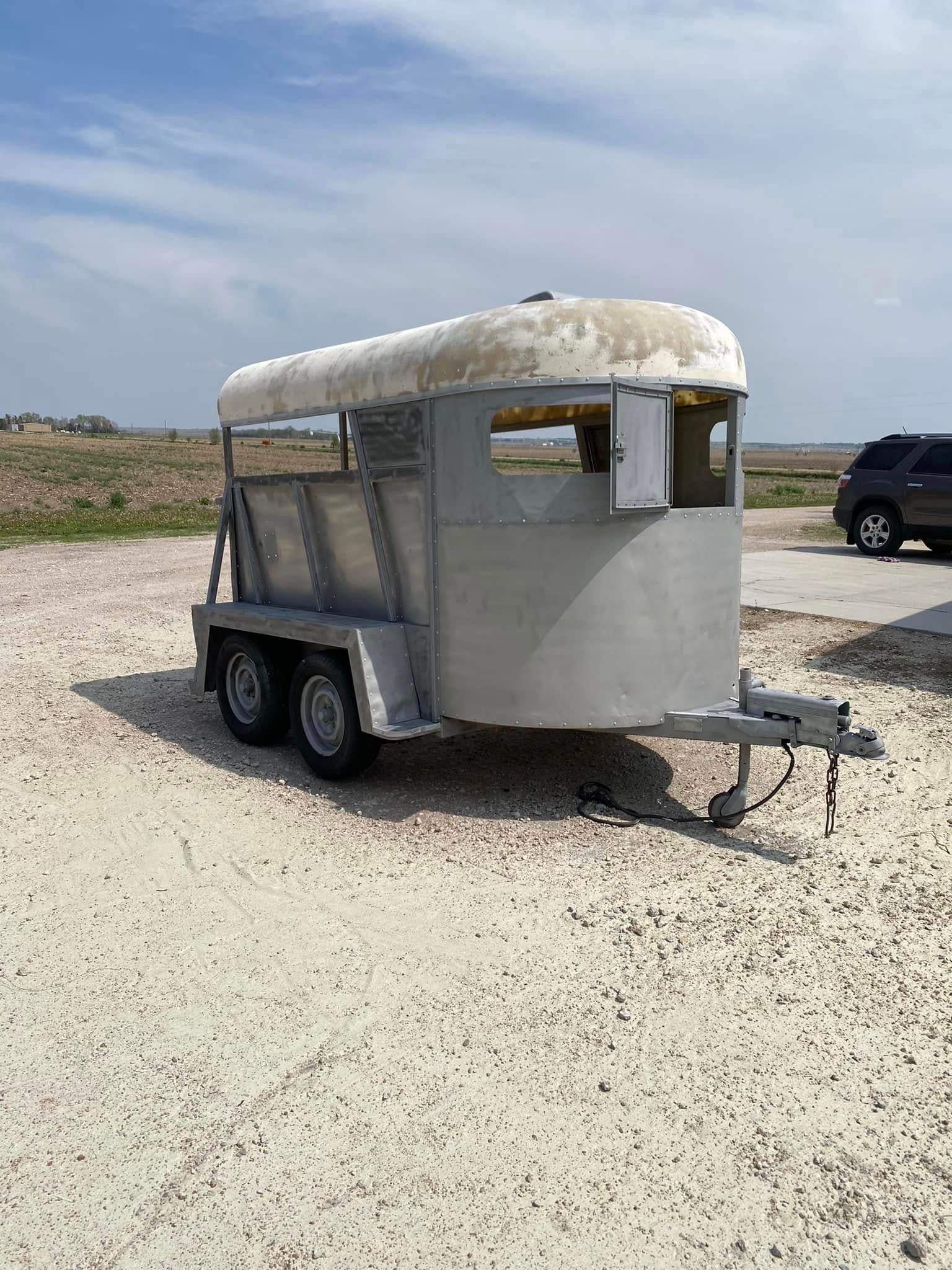 Small gray utility trailer with a white canopy parked on gravel in an open field under a cloudy sky.