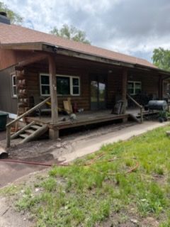 Rustic cabin porch with ramp, windows, and a grill beside grassy yard under cloudy sky