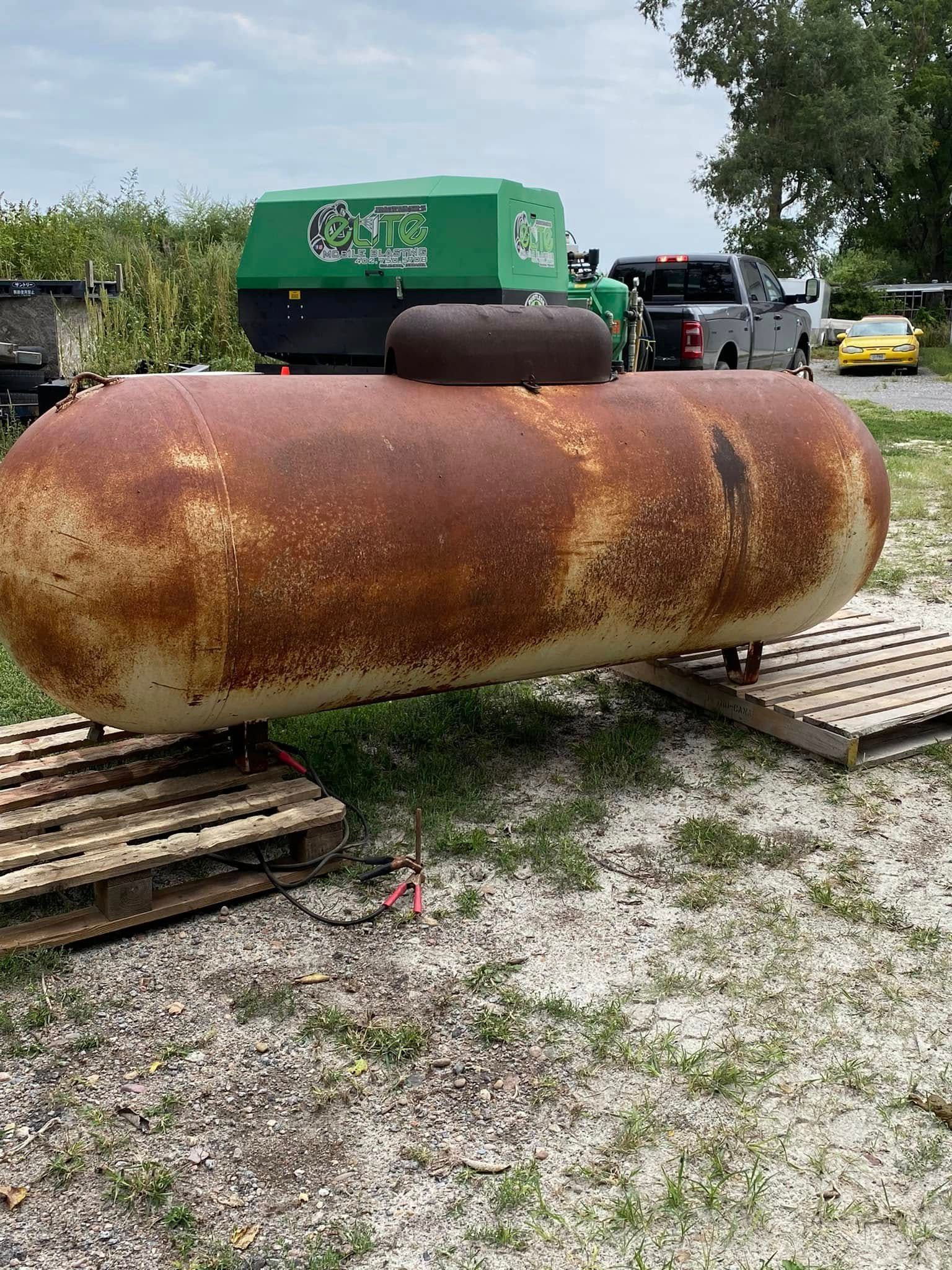 Rusty horizontal propane tank on wooden pallets in a gravel yard, with a green machine behind it.