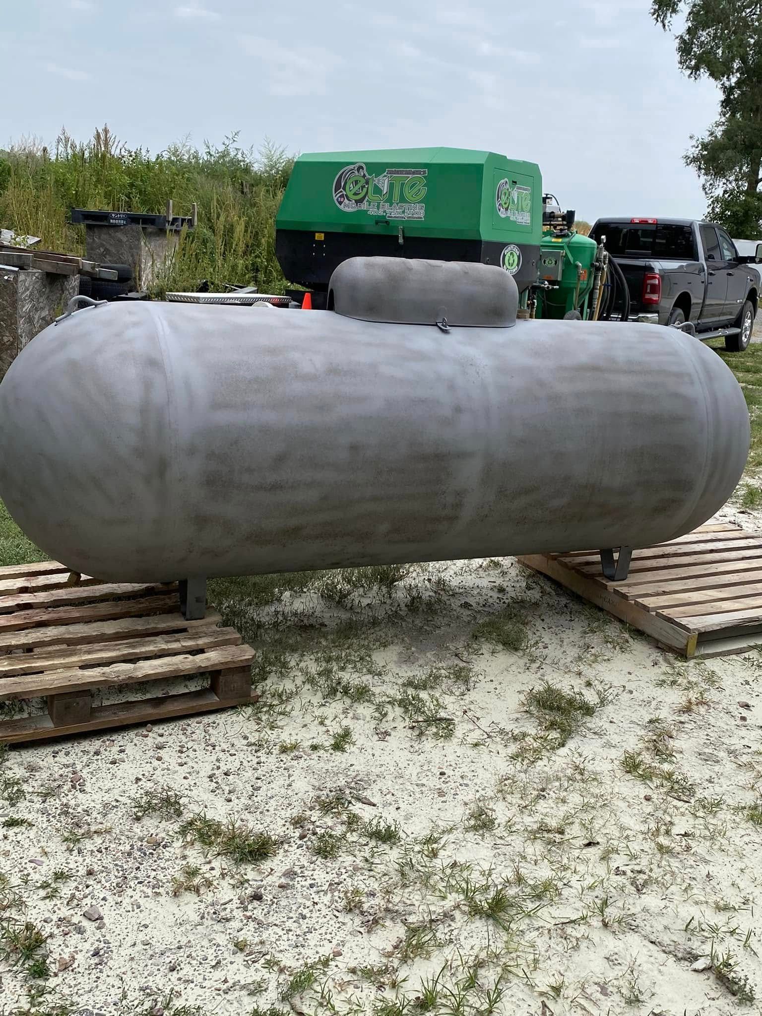 Large gray cylindrical tank on wooden pallets in an outdoor industrial yard, with a green machine behind it