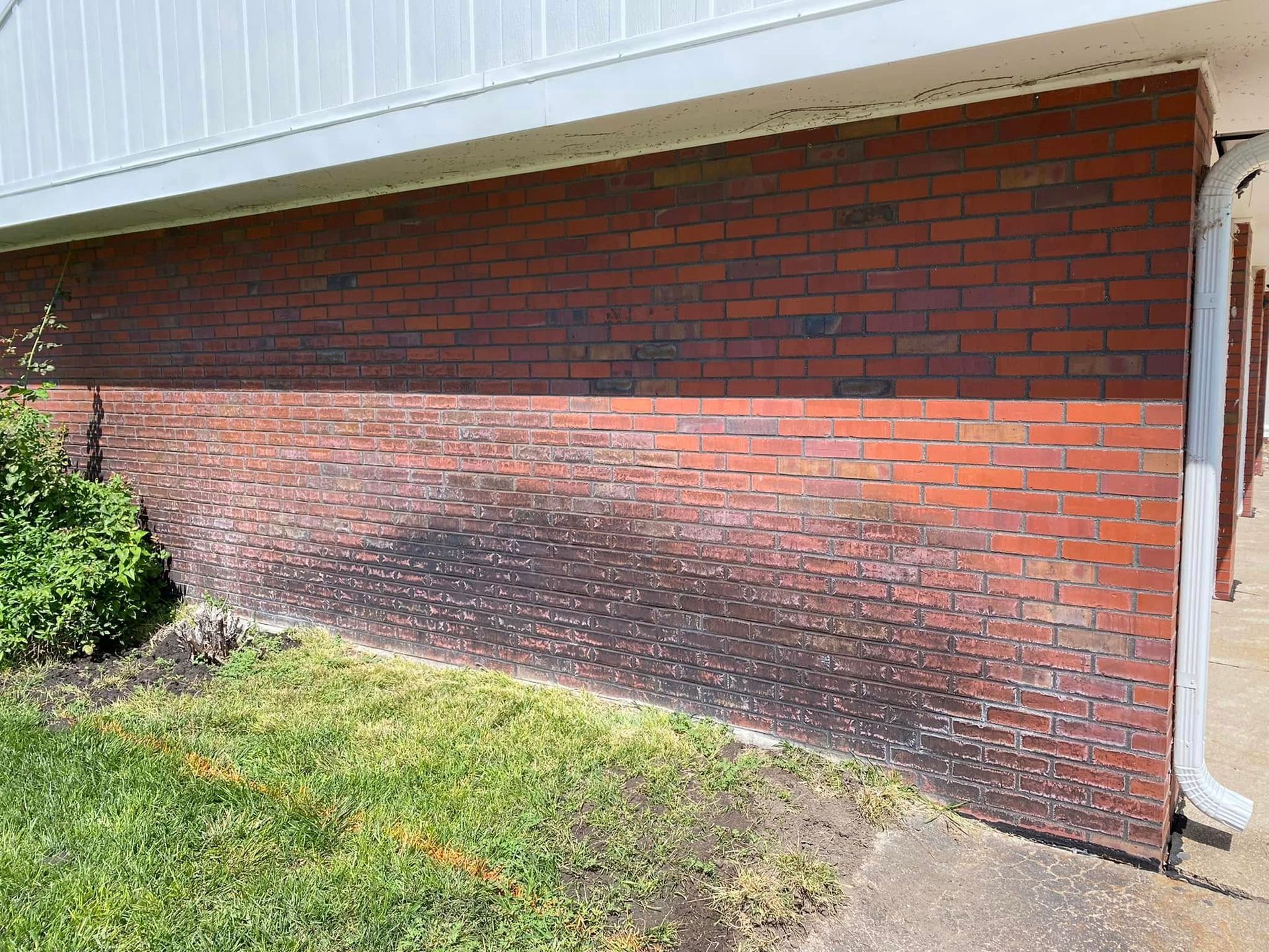 Red brick house wall with white siding, a dark stain across the lower bricks, and a small shrub at left.