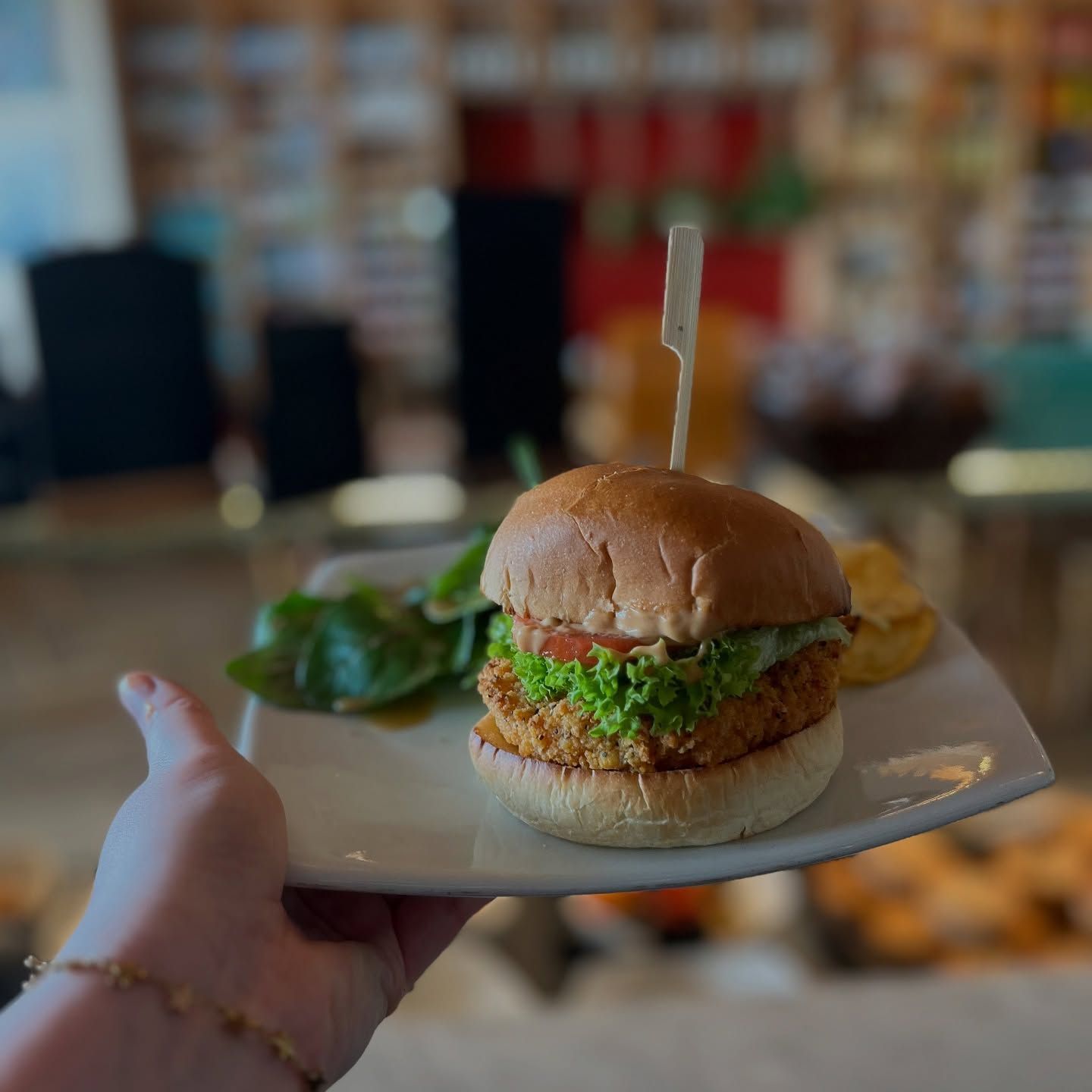 A hand holding a white plate with a small sandwich and greens in a blurred café setting