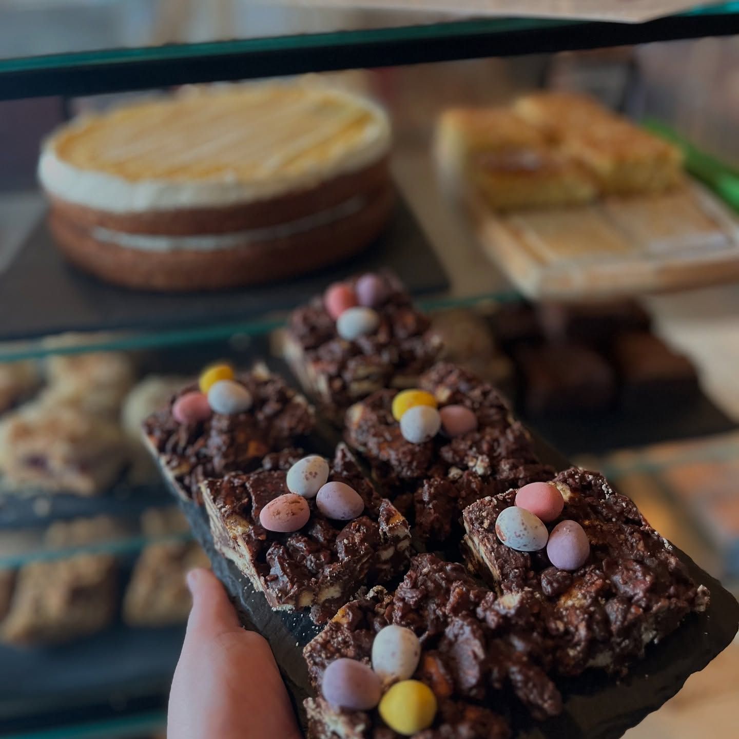 Hand holding chocolate dessert bars topped with pastel candy pieces on a bakery table