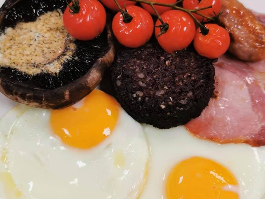 Breakfast plate with fried eggs, black pudding, bacon, sausage, mushroom, and cherry tomatoes