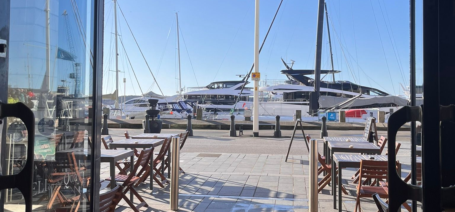 Marina view through open doors, with sailboats and a large yacht under a clear blue sky.