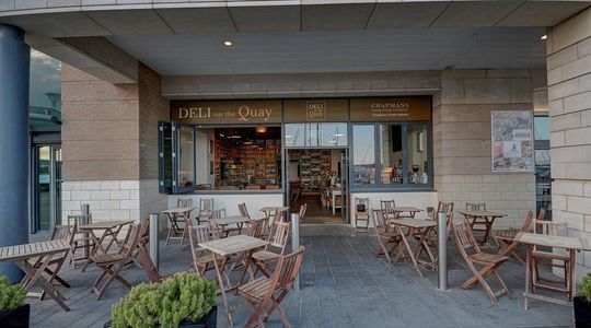 Outdoor café seating in front of a storefront, with wooden tables and chairs under a covered patio.
