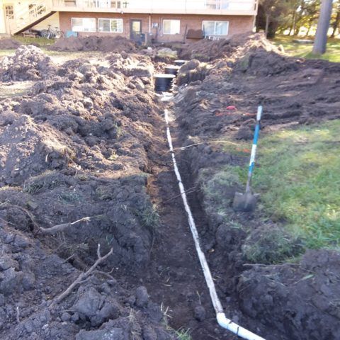 Trenched yard with a white pipe laid in the soil beside a house and grass lawn.