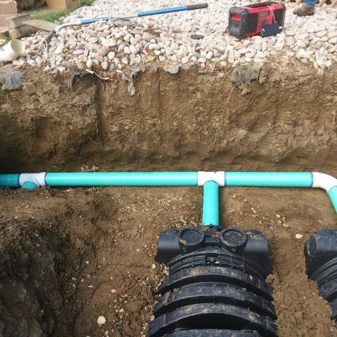 Trench with turquoise PVC pipe and black fittings beneath a gravel bed, with tools on the surface above.