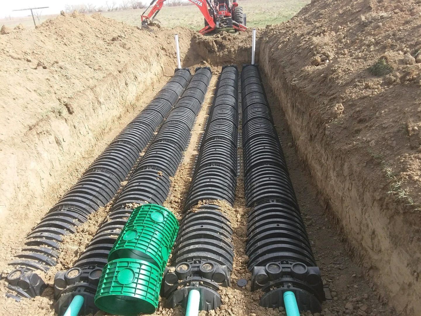 Trenches with black drainage pipes and a green pipe section being laid by a red excavator
